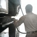 A person wearing military camouflage uniform is using a pressure washer to clean a large dark-colored military vehicle or equipment. The image is taken from behind the individual, with strong backlighting creating a silhouette effect. Water spray and debris are visibly dispersing from the point of contact with the surface being cleaned.