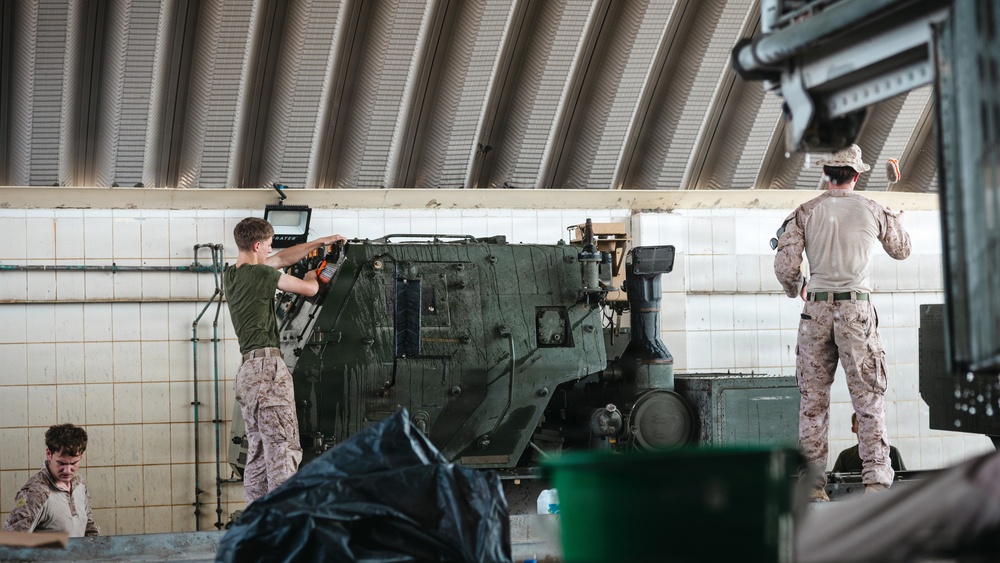 Military personnel in camouflage uniforms are performing maintenance or cleaning operations on a green armored military vehicle inside an enclosed facility. The setting appears to be a wash rack or maintenance bay with tiled walls and a corrugated metal roof. At least three service members are visible, engaged in different tasks around the vehicle.