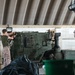 Military personnel in camouflage uniforms are performing maintenance or cleaning operations on a green armored military vehicle inside an enclosed facility. The setting appears to be a wash rack or maintenance bay with tiled walls and a corrugated metal roof. At least three service members are visible, engaged in different tasks around the vehicle.