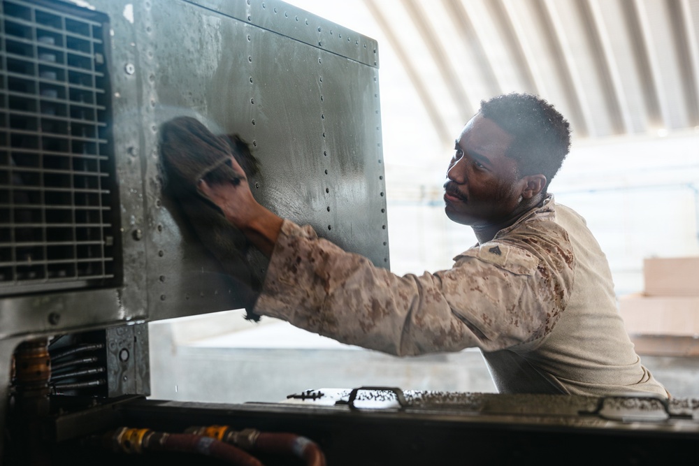 A military service member in camouflage uniform is performing maintenance work on a large piece of equipment, wiping down a metal panel with a cloth. The individual's uniform is visibly soiled with grease or oil, indicating hands-on mechanical work. The setting appears to be inside a hangar or covered maintenance facility.