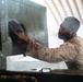 A military service member in camouflage uniform is performing maintenance work on a large piece of equipment, wiping down a metal panel with a cloth. The individual's uniform is visibly soiled with grease or oil, indicating hands-on mechanical work. The setting appears to be inside a hangar or covered maintenance facility.