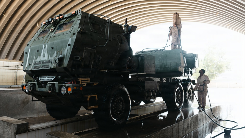 Two military personnel in camouflage uniforms are washing a large military vehicle, identified as an M142 HIMARS (High Mobility Artillery Rocket System), inside an arched maintenance facility. One soldier stands on top of the vehicle while another operates a pressure hose at ground level. The vehicle is positioned on a raised wash platform with water visibly running off the surface.
