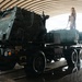 Two military personnel in camouflage uniforms are washing a large military vehicle, identified as an M142 HIMARS (High Mobility Artillery Rocket System), inside an arched maintenance facility. One soldier stands on top of the vehicle while another operates a pressure hose at ground level. The vehicle is positioned on a raised wash platform with water visibly running off the surface.