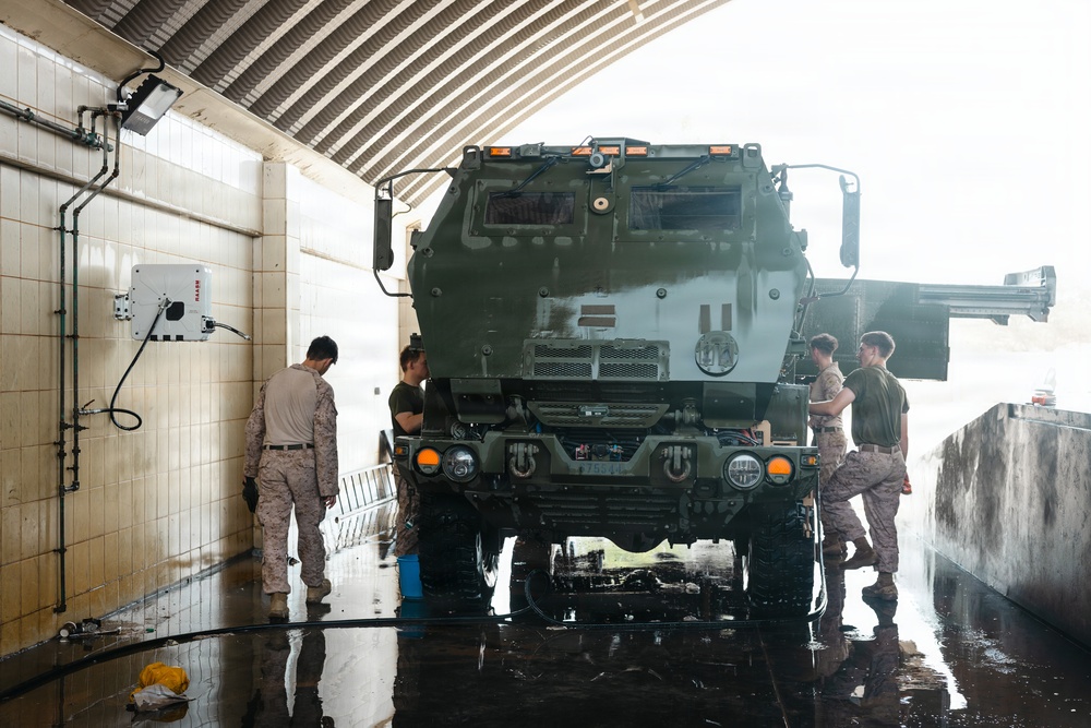 Military personnel in camouflage uniforms are washing a large olive-drab military vehicle, likely an M142 HIMARS or similar rocket artillery system, inside a covered wash bay facility. The wet concrete floor and active water hoses indicate an ongoing vehicle maintenance cleaning operation. Four service members are positioned around the front and sides of the vehicle.