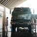 Military personnel in camouflage uniforms are washing a large olive-drab military vehicle, likely an M142 HIMARS or similar rocket artillery system, inside a covered wash bay facility. The wet concrete floor and active water hoses indicate an ongoing vehicle maintenance cleaning operation. Four service members are positioned around the front and sides of the vehicle.