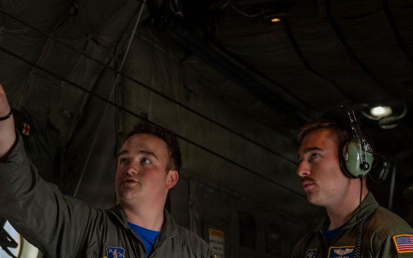 Two military personnel in olive green flight suits are inside what appears to be a military aircraft cargo bay or interior. The individual on the left is pointing upward at equipment while holding a tablet device, appearing to instruct or brief the person on the right. The person on the right is wearing aviation headphones and also holding a tablet, suggesting a training or inspection scenario.