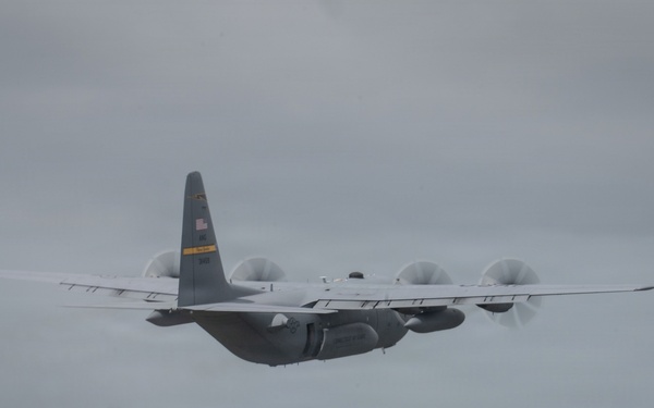 A military turboprop transport aircraft, consistent with a C-130 Hercules configuration, is photographed in flight under overcast skies. The aircraft displays an American flag on its tail fin and appears to be in a gray military paint scheme. A hazy, green landscape is visible far below.