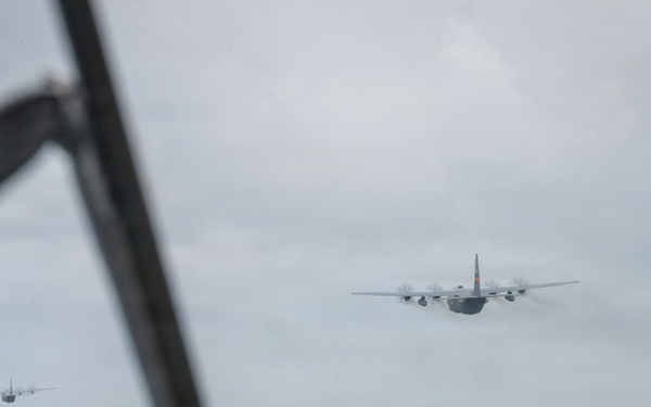 The image is taken from inside a military aircraft looking out through the windscreen or window at a formation of what appear to be C-130 Hercules turboprop transport aircraft flying in formation over a coastal or oceanic environment. At least four aircraft are visible, with one prominent in the foreground-right and two to three more visible in the mid-left distance. The sky is overcast and hazy, with a coastline or sandbar faintly visible on the horizon.