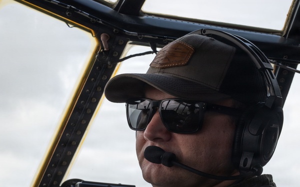 A military pilot is seated in the cockpit of an aircraft, wearing a flight suit, aviation headset with boom microphone, sunglasses, and a baseball cap. The cockpit features modern avionics including digital navigation displays and mapping screens. A second crew member is partially visible on the right side of the frame.