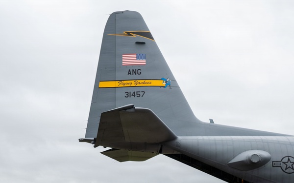 A line of fully equipped military personnel in combat gear and parachute rigs are boarding a U.S. Air National Guard C-130 Hercules transport aircraft. The aircraft's tail markings identify it as belonging to the 'Flying Yankees,' tail number 31457, of the Air National Guard (ANG). The scene appears to be a parachute operation or airborne exercise conducted on a grass airfield.