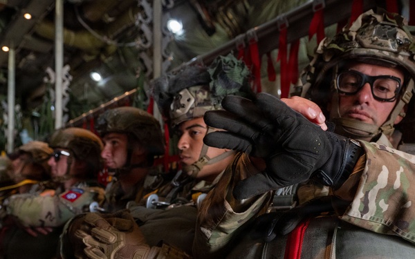 A group of military paratroopers are seated inside a military transport aircraft, fully equipped with parachute packs, helmets, tactical gloves, and combat gear. The soldiers appear to be preparing for an airborne jump operation, with red static line straps visible overhead. The foreground soldier is wearing protective goggles and adjusting equipment while seated in a row with fellow paratroopers.