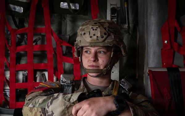 A military service member in full combat gear is seated inside what appears to be a military transport aircraft, likely a C-130 or similar cargo plane. The soldier is wearing multicam/OCP pattern uniform, a tactical helmet with NVG mount, body armor, and a parachute harness, suggesting preparation for an airborne operation. The individual appears focused and contemplative, gazing to the side.