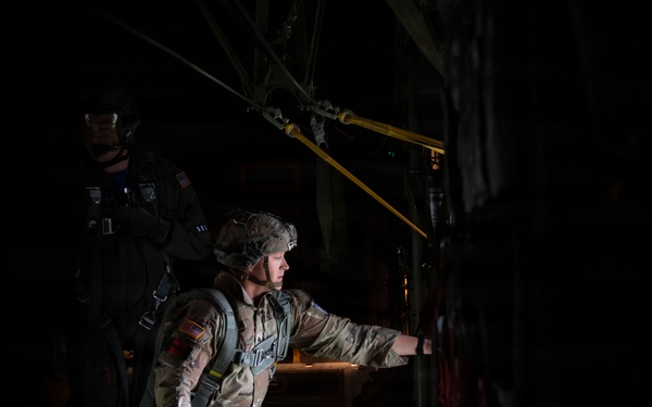 A U.S. Army soldier in full combat gear and parachute harness stands near the open door or ramp of a military transport aircraft, appearing to be preparing for or conducting an airborne operation. The image is dramatically lit with most of the frame in deep shadow, highlighting the soldier's face and equipment. A second soldier is partially visible in the background.