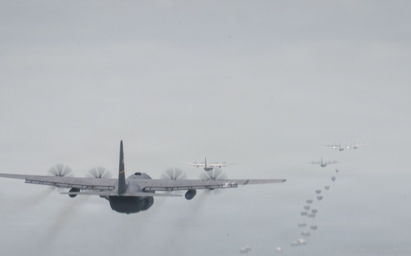 Multiple military transport aircraft, consistent with C-130 Hercules variants, are flying in formation while conducting an airdrop operation. Small parachute-deployed objects are visibly falling from the aircraft in a trail pattern below and behind the formation. The image is taken from an aerial perspective, likely from another aircraft, over a hazy landscape.