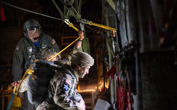 Military personnel are conducting a static-line parachute jump from the rear ramp of a military transport aircraft, likely a C-130 Hercules. A jumpmaster in flight crew gear is visible holding the static line cable while a paratrooper in full combat gear and parachute rig prepares to exit the aircraft. The scene is dramatically lit with the dark interior of the aircraft contrasting against the open ramp.