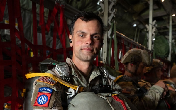 A U.S. Army soldier wearing 82nd Airborne Division insignia is seated inside a military transport aircraft, equipped with full parachute gear. He is posed facing the camera with a calm expression while other soldiers are visible seated behind him. The setting indicates a pre-jump airborne operation.