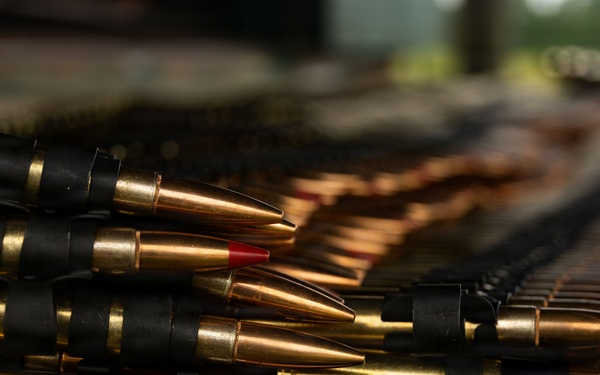 The image shows multiple rows of ammunition rounds secured in black metal belt links, forming machine gun ammunition belts. The cartridges have brass casings with pointed projectiles, and some rounds display red-tipped bullets, indicating a specific ammunition type such as tracer or incendiary rounds. The belts extend into the background with depth-of-field blur.