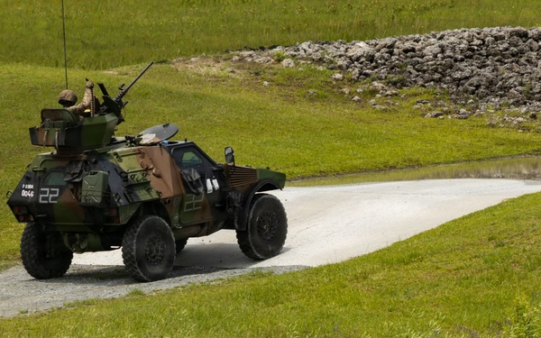 A military armored vehicle is traveling along a paved/gravel road through a green grassy field. The vehicle appears to be a light armored reconnaissance vehicle with a mounted weapon system and at least one soldier visible in the turret. The setting appears to be a military training area or exercise ground.