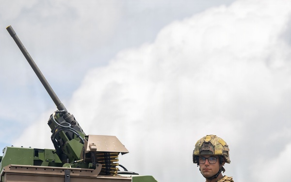 A soldier in full combat gear is positioned in the turret hatch of an armored military vehicle, looking directly at the camera. The vehicle is painted in a tan and green camouflage scheme and is equipped with a remote weapon station or autocannon mounted on top. The image appears to be taken during a military exercise or demonstration in an outdoor setting.