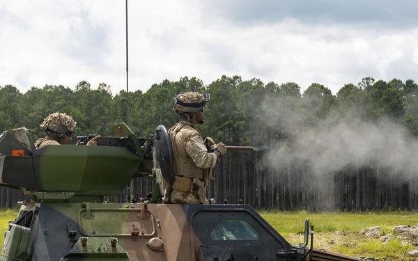 Two soldiers are operating a light armored military vehicle marked with the number '22' in a green and brown camouflage pattern. A weapon system appears to have just been fired, as evidenced by visible smoke/exhaust emanating from the right side of the vehicle. The scene takes place on an open grassy field with a tree line in the background, consistent with a military training range.