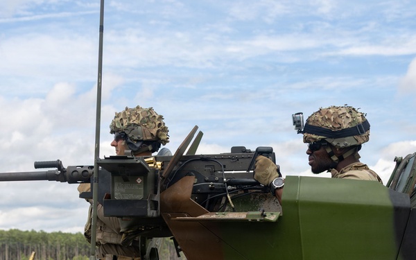 Two military personnel are positioned in the turret of an armored vehicle, equipped with a mounted weapon system. The soldiers are wearing camouflage helmets and tactical gear, operating in an open field environment with tree lines and water visible in the background. The scene appears to depict a training exercise or operational deployment.