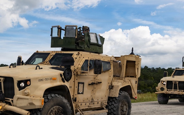 Two tan/desert-colored Joint Light Tactical Vehicles (JLTVs) are positioned on a gravel road or staging area, marked with USMC identification numbers. Both vehicles are equipped with turret-mounted weapon systems and appear to be in an operational or training configuration. The setting appears to be a rural training environment with tree lines visible in the background.