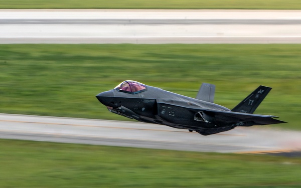 A dark-colored fifth-generation stealth fighter jet, consistent with an F-35 Lightning II, is captured at low altitude just above a runway during takeoff or a high-speed pass. The image uses a panning photography technique, creating motion blur in the background while keeping the aircraft relatively sharp. The aircraft appears to carry unit markings on its tail fin.