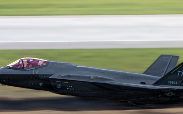 A panning-shot photograph captures an F-35A Lightning II fighter jet at low altitude during a high-speed takeoff or low pass over a runway. The tail code 'TY' and serial number '5804' are visible, indicating assignment to Tyndall Air Force Base. A red pickup truck is visible in the foreground, also motion-blurred, providing scale and context to the aircraft's speed.