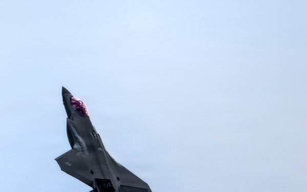 A dark-colored fifth-generation stealth fighter jet, consistent with an F-35 Lightning II, is captured in a steep climbing maneuver against a pale blue sky. The aircraft's single engine is visibly producing afterburner thrust, with an orange flame and white exhaust plume trailing behind. The canopy appears to have a reddish-purple tint, typical of modern fighter jet canopy coatings.