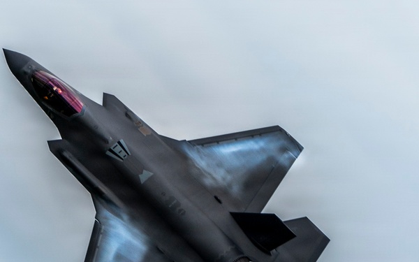 A dark-colored fifth-generation stealth fighter jet is photographed from above at a steep angle, appearing to perform a high-performance maneuver. The aircraft's single engine is visibly lit with afterburner flame, indicating high thrust output. The overall composition and aircraft design are consistent with an F-35 Lightning II.
