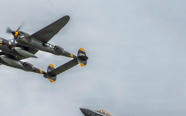 Two military aircraft are flying in formation against an overcast sky, representing approximately 80 years of aviation advancement. The upper aircraft is a World War II-era Lockheed P-38 Lightning with twin-boom configuration, marked with the number 162 and U.S. star insignia. The lower aircraft is a modern Lockheed Martin F-35 Lightning II stealth fighter jet, creating a deliberate heritage formation pairing of the two 'Lightning' aircraft.