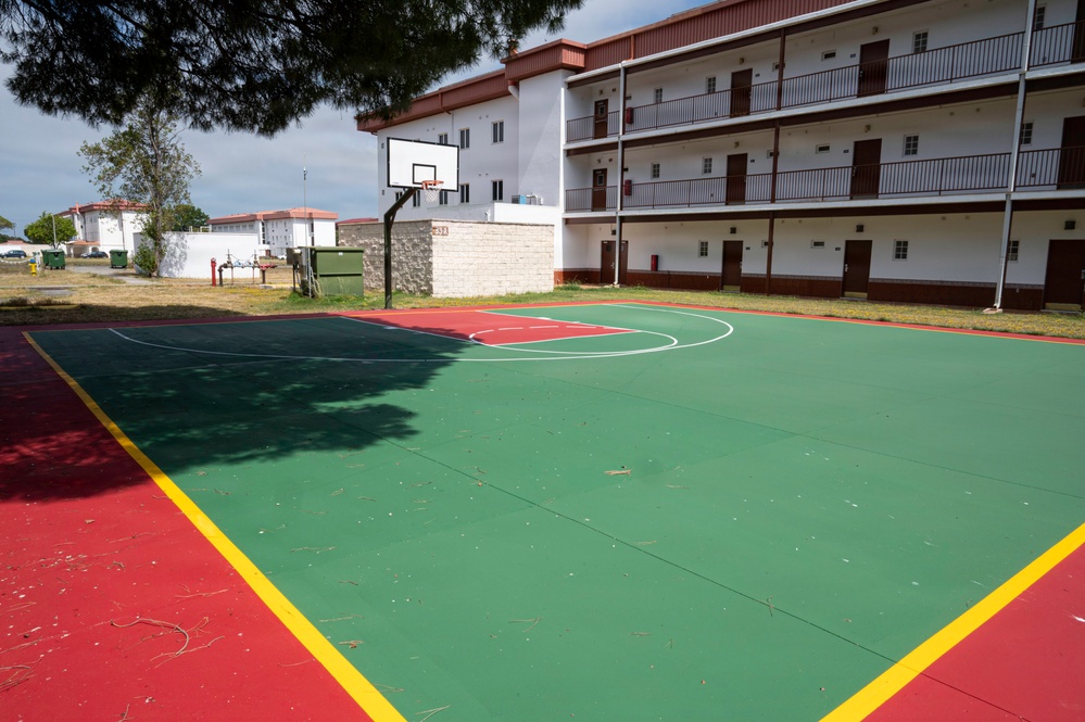An outdoor basketball court with a green and red surface, outlined by yellow boundary lines, situated adjacent to a multi-story white building with red trim and balconies. The court features a single visible basketball hoop mounted on a stone/brick base structure. The area appears to be part of an institutional or residential complex, possibly a military barracks or dormitory facility.