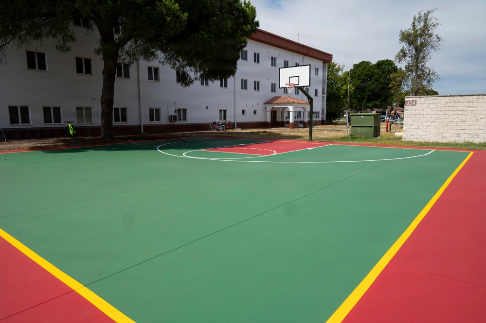 An outdoor basketball court with a green and red surface outlined by yellow boundary lines, located adjacent to a multi-story white building with a red roof. The court features a single basketball hoop with backboard mounted on a metal pole at the far end. The area appears to be part of a residential or institutional complex, with trees, bicycles, and additional structures visible in the background.