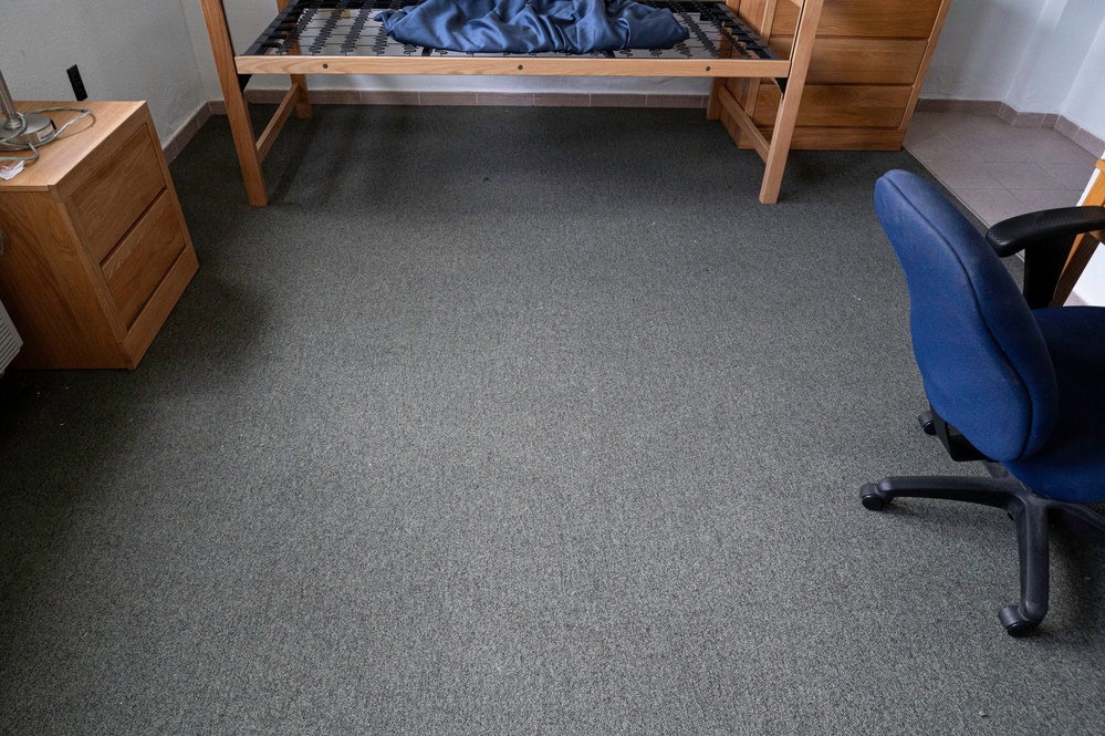 The image shows a sparsely furnished bedroom, likely a dormitory or student room, photographed from above at an angle. The room contains a lofted wooden bed frame without a mattress, a wooden nightstand, and a blue office chair. The floor is covered with gray carpet and the room appears to be in a partially moved-in or moved-out state.