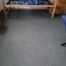 The image shows a sparsely furnished bedroom, likely a dormitory or student room, photographed from above at an angle. The room contains a lofted wooden bed frame without a mattress, a wooden nightstand, and a blue office chair. The floor is covered with gray carpet and the room appears to be in a partially moved-in or moved-out state.