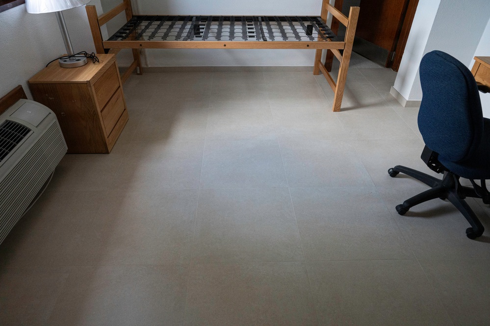 A sparse, minimally furnished room with a tile floor, featuring a bed frame without a mattress, a wooden nightstand, and a blue office chair. The room appears to be in a transitional state, possibly a dormitory or budget accommodation being set up or cleared out. Notable is the absence of bedding or a mattress on the slatted bed frame.