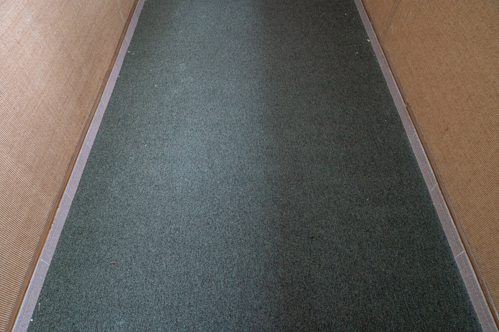 The image shows a narrow corridor or hallway viewed from above at a low angle, featuring a dark green carpet runner laid along the center. The runner is flanked on both sides by lighter tan or beige textured flooring material, separated by thin metal or plastic edging strips. The perspective creates a converging vanishing point effect toward the far end of the passage.