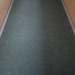 The image shows a narrow corridor or hallway viewed from above at a low angle, featuring a dark green carpet runner laid along the center. The runner is flanked on both sides by lighter tan or beige textured flooring material, separated by thin metal or plastic edging strips. The perspective creates a converging vanishing point effect toward the far end of the passage.
