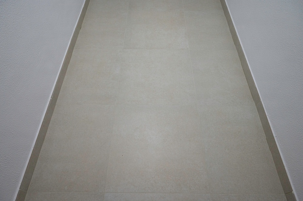 The image shows a narrow corridor or hallway viewed from a low angle looking down its length. The floor is covered with large, light beige/cream ceramic or porcelain tiles with minimal grout lines visible. The walls on both sides are white with a textured finish, and a thin border trim separates the wall from the floor.