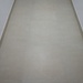 The image shows a narrow corridor or hallway viewed from a low angle looking down its length. The floor is covered with large, light beige/cream ceramic or porcelain tiles with minimal grout lines visible. The walls on both sides are white with a textured finish, and a thin border trim separates the wall from the floor.