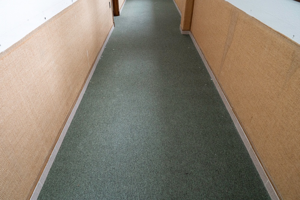 The image shows a narrow, empty interior hallway with a dark green carpet runner extending toward a doorway in the background. The walls are lined with tan or beige textured paneling, and the upper portions of the walls are white. The corridor appears institutional or utilitarian in nature, with no people or furnishings present.