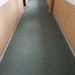 The image shows a narrow, empty interior hallway with a dark green carpet runner extending toward a doorway in the background. The walls are lined with tan or beige textured paneling, and the upper portions of the walls are white. The corridor appears institutional or utilitarian in nature, with no people or furnishings present.