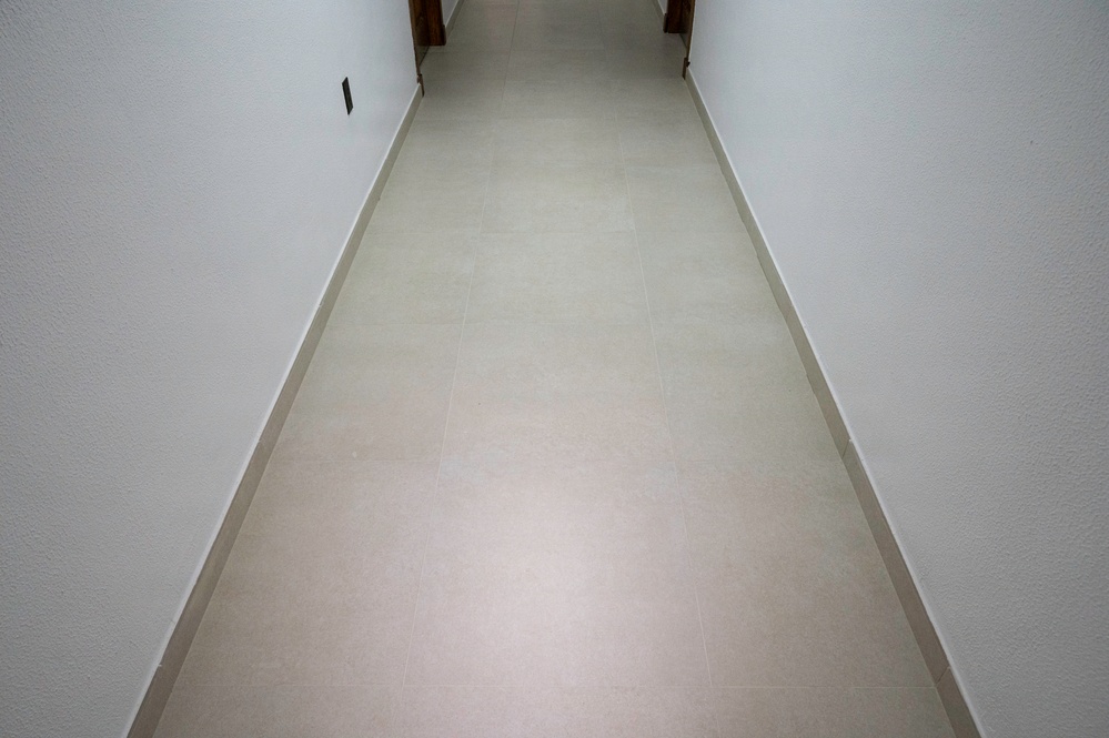 The image shows a narrow residential hallway with light-colored ceramic or porcelain tile flooring and white painted walls. The corridor leads toward a wooden door visible at the far end. The space appears clean, modern, and unoccupied.