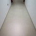 The image shows a narrow residential hallway with light-colored ceramic or porcelain tile flooring and white painted walls. The corridor leads toward a wooden door visible at the far end. The space appears clean, modern, and unoccupied.
