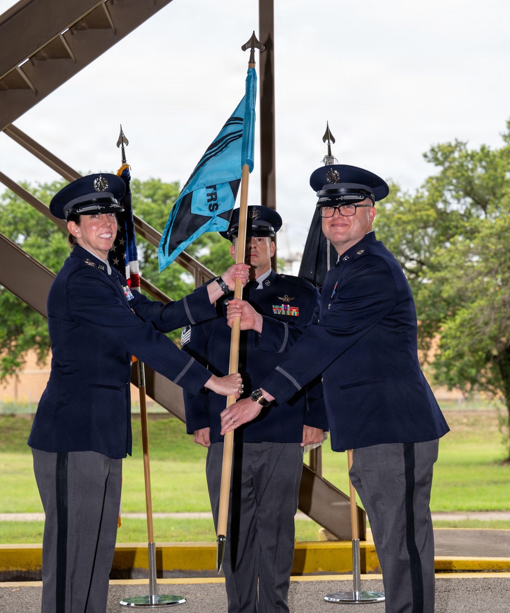 A military change of command ceremony is taking place outdoors, likely at a U.S. Air Force installation. Two officers are exchanging a light blue guidon flag marked 'TRS' while a senior non-commissioned officer stands between them holding the staff. The ceremony follows standard U.S. Air Force protocol for unit command transitions.