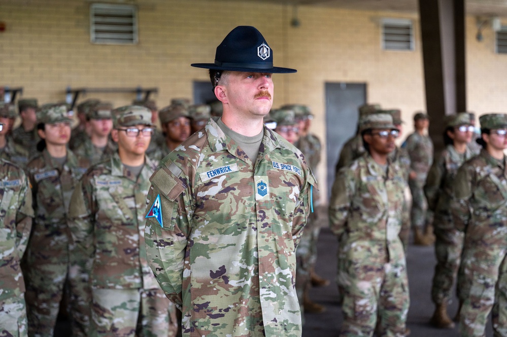 A U.S. Space Force Military Training Instructor (MTI) wearing a distinctive black campaign/drill sergeant hat stands at attention in the foreground, identified by the name tape 'FENWRICK' and 'U.S. SPACE FORCE' tape on his OCP uniform. He appears to be overseeing a formation of trainees wearing standard OCP camouflage uniforms and patrol caps in the background. The setting appears to be a covered outdoor drill pad or bay at a military training installation.