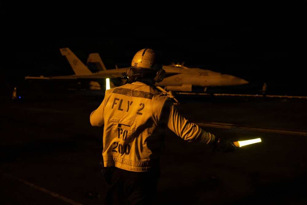 A night flight deck operation aboard what appears to be a U.S. Navy aircraft carrier. A flight deck crew member wearing a yellow jersey marked 'FLY 2 / PO / 200' is directing aircraft using a luminescent wand. A military jet aircraft, visible with the number '305' on its fuselage and consistent with an F/A-18 Hornet or Super Hornet, is positioned on the deck in the background.
