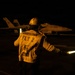 A night flight deck operation aboard what appears to be a U.S. Navy aircraft carrier. A flight deck crew member wearing a yellow jersey marked 'FLY 2 / PO / 200' is directing aircraft using a luminescent wand. A military jet aircraft, visible with the number '305' on its fuselage and consistent with an F/A-18 Hornet or Super Hornet, is positioned on the deck in the background.
