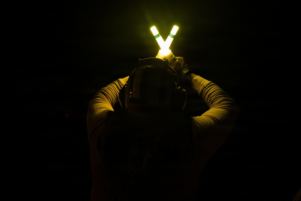 A person photographed from behind in near-total darkness is holding two illuminated wands or light sticks above their head in a V-shape configuration. The bright yellow-white glow of the wands with green banding is the primary light source in the image, casting a faint warm illumination on the person's upper body. The individual appears to be wearing tactical or military-style gear including a helmet and long-sleeved shirt.