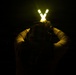 A person photographed from behind in near-total darkness is holding two illuminated wands or light sticks above their head in a V-shape configuration. The bright yellow-white glow of the wands with green banding is the primary light source in the image, casting a faint warm illumination on the person's upper body. The individual appears to be wearing tactical or military-style gear including a helmet and long-sleeved shirt.